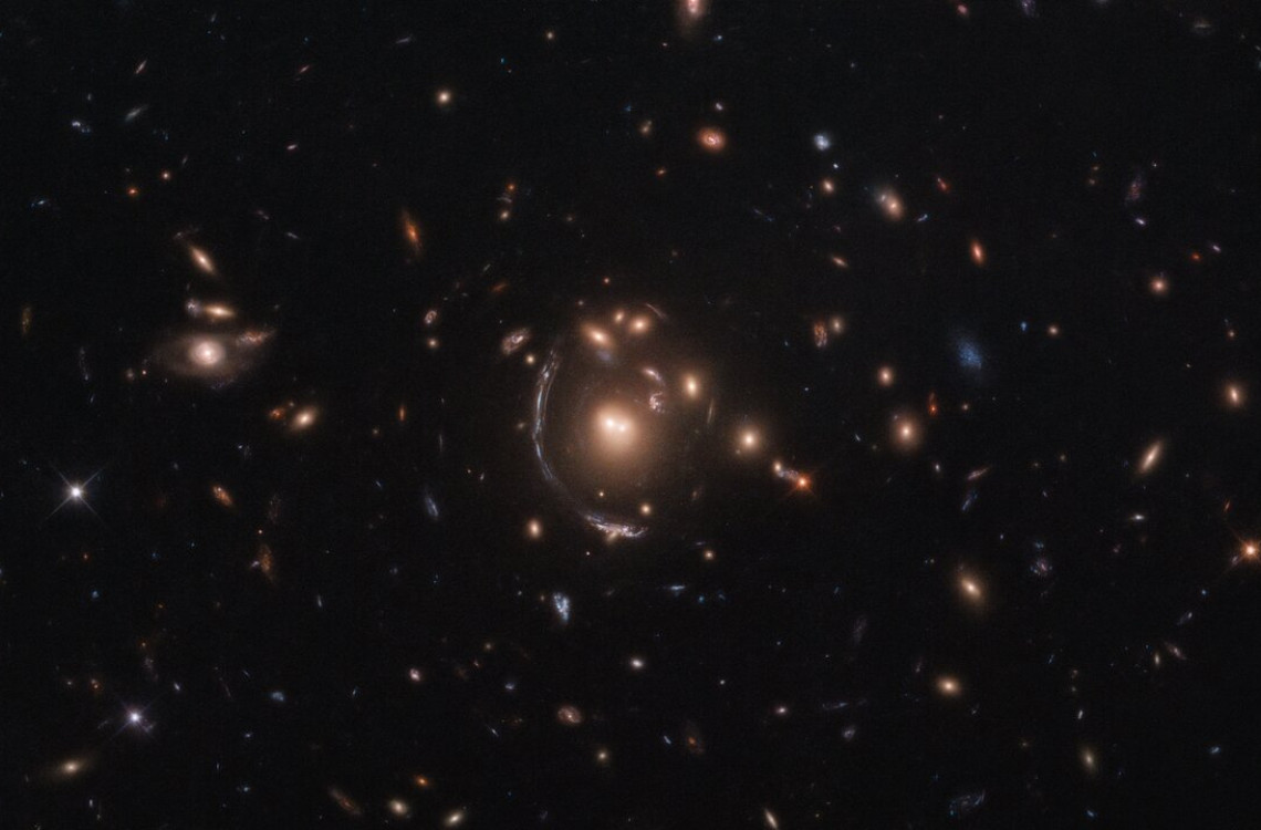 This NASA/ESA Hubble Space Telescope image features the galaxy LRG-3-817, also known as SDSS J090122.37+181432.3. The galaxy, its image distorted by the effects of gravitational lensing, appears as a long arc to the left of the central galaxy cluster. Gravitational lensing occurs when a large distribution of matter, such as a galaxy cluster, sits between Earth and a distant light source. As space is warped by massive objects, the light from the distant object bends as it travels to us and we see a distorted image of it. This effect was first predicted by Einstein’s general theory of relativity. Strong gravitational lenses provide an opportunity for studying properties of distant galaxies, since Hubble can resolve details within the multiple arcs that are one of the main results of gravitational lensing. An important consequence of lensing distortion is magnification, allowing us to observe objects that would otherwise be too far away and too faint to be seen. Hubble makes use of this magnification effect to study objects beyond the sensitivity of its 2.4-metre-diameter primary mirror, showing us the most distant galaxies humanity has ever encountered. This lensed galaxy was found as part of the Sloan Bright Arcs Survey, which discovered some of the brightest gravitationally lensed high-redshift galaxies in the night sky. 